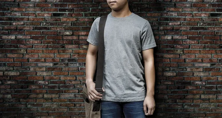 Man wearing a plain grey crew-neck T-shirt and messenger bag against a brick wall—representing everyday apparel and responsible textile sourcing.