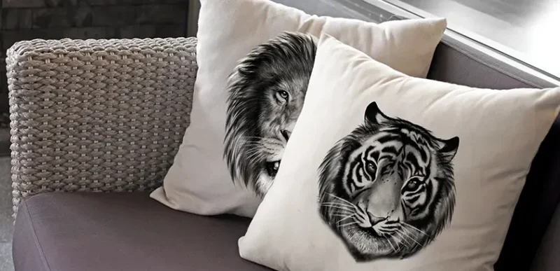 Close-up of two custom-printed cushion covers on a cane outdoor sofa—one with a black-and-white tiger print, one with a lion—showing detail of fabric and print quality.