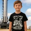Boy wearing a black I Really Need More Space Kids T Shirt standing in front of a small rocket