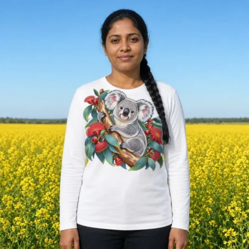 Woman with a long black plait over one shoulder beside a bright yellow canola field under a blue sky, wearing the white Koala in Coral Eucalyptus women’s long sleeve T-shirt