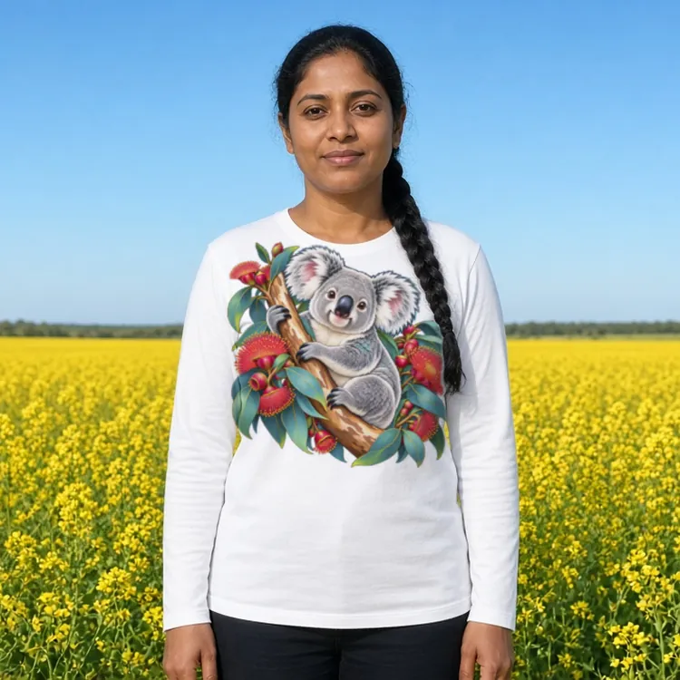 Woman with a long black plait over one shoulder beside a bright yellow canola field under a blue sky, wearing the white Koala in Coral Eucalyptus women’s long sleeve T-shirt