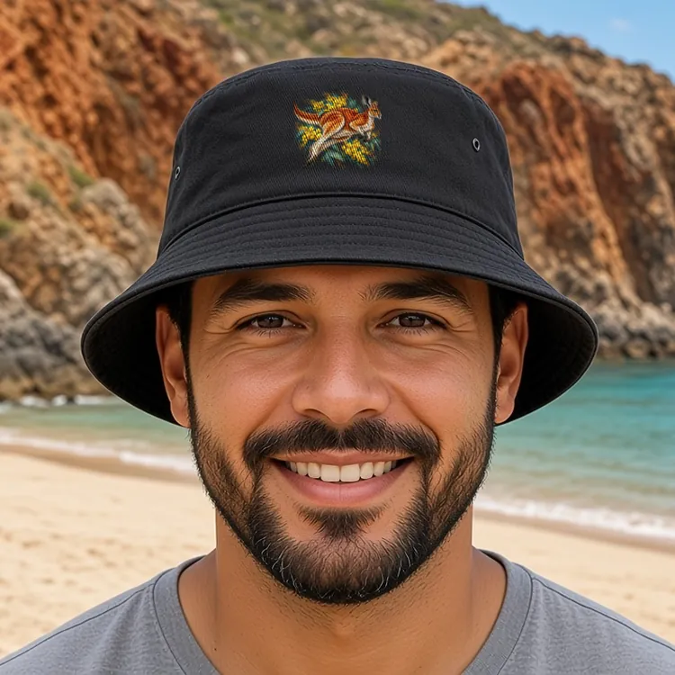 Smiling man with a short dark beard on a beach below red rock cliffs, face to camera, wearing the black Red Kangaroo and Golden Wattle bucket hat