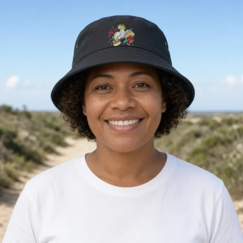Smiling woman with short curly brown hair on a sandy track through coastal heath and banksias, face to camera, wearing the black Sulphur-crested Cockatoo in Bloom bucket hat
