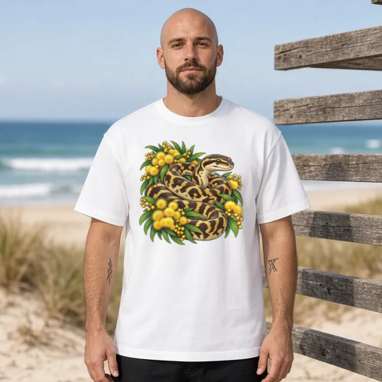 Man with a shaved head and short beard beside a weathered timber rack near the beach, wearing the white Carpet Python Among Golden Wattle boxy T-shirt