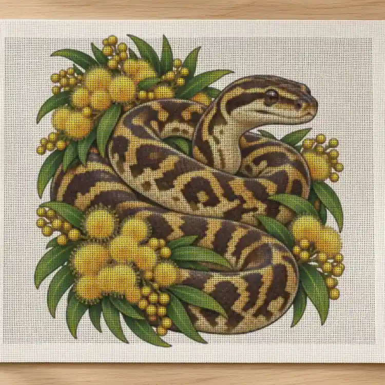 Carpet Python Among Golden Wattle cross stitch canvas on a timber craft table with yellow, green and brown threads and scissors